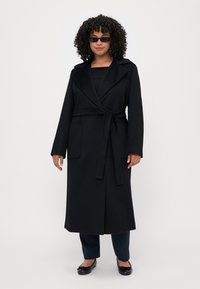 Cappotto lungo nero con vita fasciata, ampio colletto e due tasche laterali, realizzato in tessuto morbido, abbinato a pantaloni scuri e ballerine.