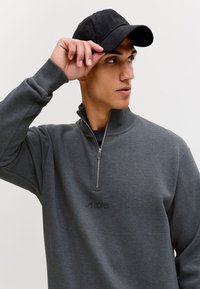 Junger Mann, der eine schwarze Kappe richtet, trägt ein dunkelgraues Sweatshirt mit Viertelreißverschluss und hochgestelltem Kragen vor einem schlichten hellen Hintergrund.