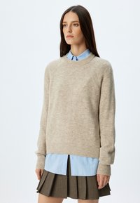 Maglione beige a maglia con polsini e orlo a coste, indossato sopra una camicia colletto azzurro chiaro. Modello abbinato a una gonna marrone plissettata.