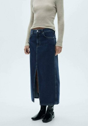 Gonna di jeans, blu scuro, lunghezza lunga con uno spacco frontale. Presenta cinque tasche e una chiusura con bottone. Abbinata a stivaletti neri aderenti.