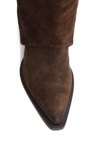 Brązowy zamszowy but z ostrym noskiem, teksturowaną górą i płaską, ciemną podeszwą. Projekt jest elegancki, z minimalnym przeszyciem i bez widocznych elementów metalowych.