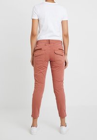 Kvinna klädd i rostfärgade slim-byxor med bakfickor och vita sneakers, stående mot en enfärgad vit bakgrund.