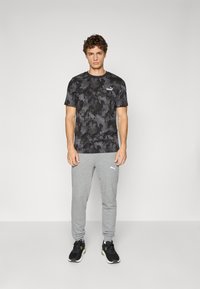 Zwarte sportieve t-shirt met een subtiel camouflagepatroon, gecombineerd met lichtgrijze joggingsbroek met een zijlogo. Zwarte sneakers maken de look compleet.