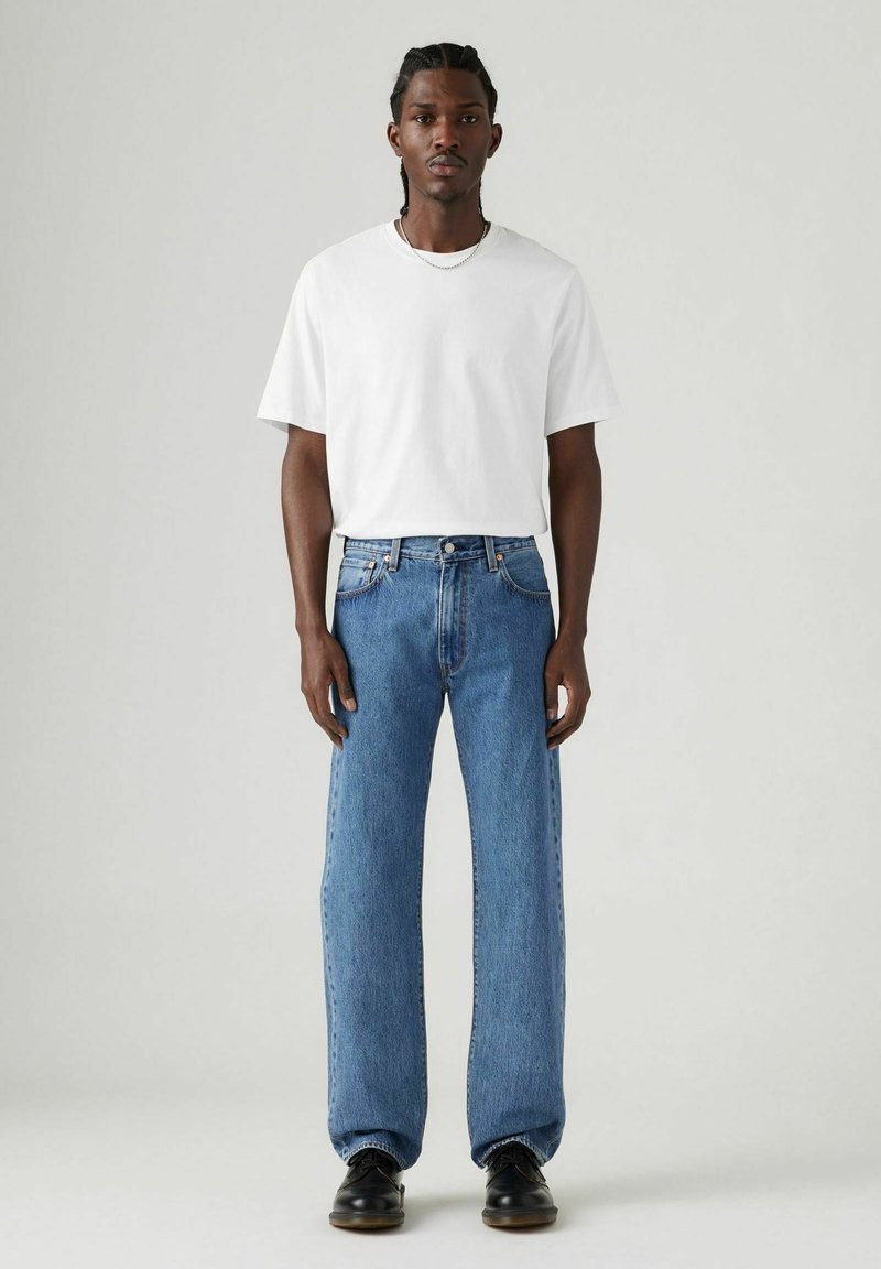 Camiseta blanca de cuello redondo combinada con vaqueros holgados de color azul claro. El modelo lleva zapatos negros. Look sencillo y casual con acentos mínimos.