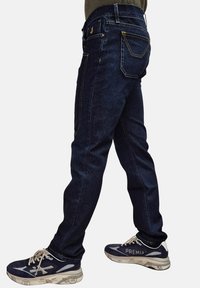 Jeans in denim blu scuro con vestibilità dritta, caratterizzati da cuciture gialle, cinque tasche e vita media. Abbinati a sneaker scure.