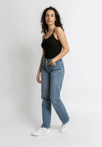 Zwarte getailleerde tanktop, lichtblauwe high-waisted jeans en witte sneakers. Model staat voor een neutrale achtergrond en toont casual kleding.