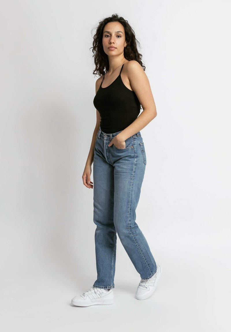 Zwarte getailleerde tanktop, lichtblauwe high-waisted jeans en witte sneakers. Model staat voor een neutrale achtergrond en toont casual kleding.
