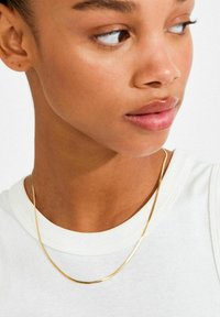 Guldhalskæde, fladt design, let vægt, poleret finish, diskret bue, båret over en hvid skjorte, hvilende på kravebensområdet.