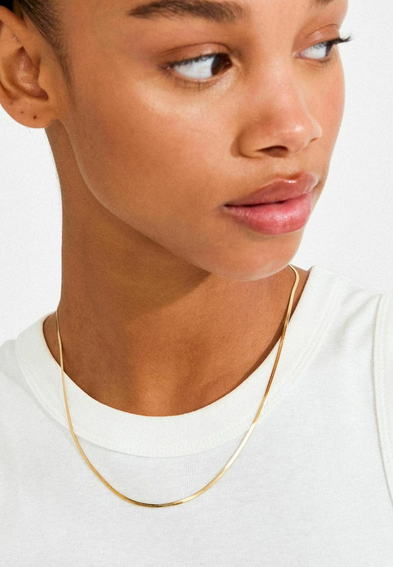 Guldhalskæde, fladt design, let vægt, poleret finish, diskret bue, båret over en hvid skjorte, hvilende på kravebensområdet.