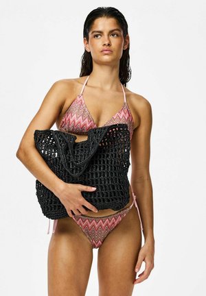 Femeie în bikini roz cu model, ținând o geantă mare neagră țesută, pe fundal alb.