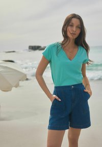 T-shirt turquoise à col en V associé à un short en denim bleu taille haute. Le short possède des poches avant et une coupe décontractée. Cadre de plage.