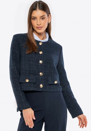 Blazer de tweed azul marino con botones dorados, diseño corto, tejido texturizado y dos bolsillos delanteros. Mangas largas con detalles en los puños.