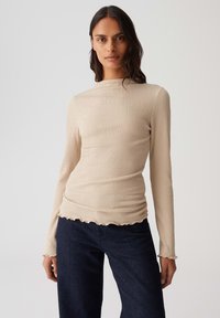 Frau in einem beigefarbenen, gerippten Langarmoberteil mit gewellten Säumen und dunkelblauen Jeans, die vor einem schlichten, hellen Hintergrund steht.