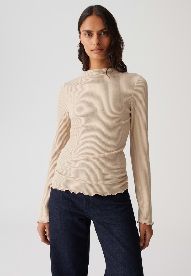 Vrouw draagt een beige ribbel top met lange mouwen en gescheurde randen, en een donkere spijkerbroek, staand tegen een effen lichte achtergrond.