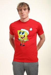 Camiseta corta roja con dibujos que presenta a un personaje de dibujos animados usando un sombrero de Santa, con una expresión sonriente y sosteniendo cajas cuadradas marrones.