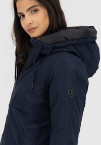 Giubbotto con cappuccio blu navy, con zip frontale, tessuto esterno testurizzato e foderatura interna liscia. Presenta un'emblema sulla manica.