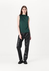 Groene satijnen mouwloze top met een gedrapeerde halslijn, gecombineerd met hoge zwarte faux leren broek en zwarte schoenen, staand op een effen achtergrond.