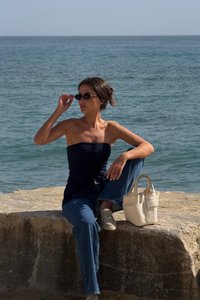 Model trägt ein navyfarbenes trägerloses Oberteil und hellblaue weit geschnittene Jeans, sitzt auf einer Steinoberfläche mit einer weißen Handtasche und schwarzen Sonnenbrillen.