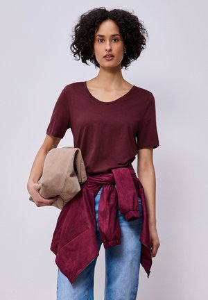 Mujer joven con cabello rizado, lleva una camiseta granate y una chaqueta anudada alrededor de la cintura, sosteniendo una chaqueta beige doblada en su brazo izquierdo.