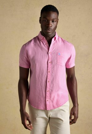 Tânăr care poartă o cămașă roz cu mâneci scurte și nasturi și pantaloni bej deschis, stând în picioare în fața unui fundal simplu bej.