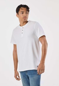 Valkoinen lyhythihainen henley-paita ribbineuloksella, jossa on kolme nappia ja pyöreä kaula-aukko. Yhdistetty sinisiin farkkuihin.