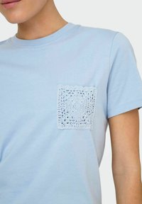 Lys blå bomulds t-shirt med en dekorativ hæklet lomme. Korte ærmer og rund halsudskæring med en glat tekstur.