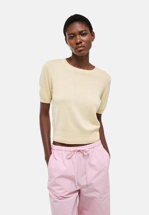 Femme portant un pull beige à manches courtes et un pantalon à cordon de serrage rose clair, debout les mains dans les poches sur un fond blanc.
