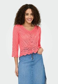 Pull en tricot rose avec un col en V, des manches 3/4 et un ourlet festonné ; présente un motif ajouré et une finition texturée. Associé à une jupe en denim.