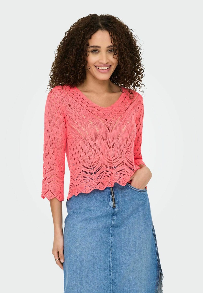 Pull en tricot rose avec un col en V, des manches 3/4 et un ourlet festonné ; présente un motif ajouré et une finition texturée. Associé à une jupe en denim.