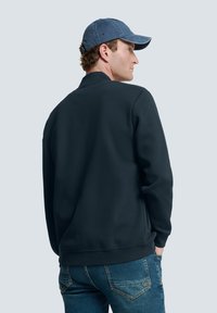 Dunkelblaues Sweatshirt mit Rundhalsausschnitt, gerippten Bündchen und Saum, kombiniert mit hellblauen Jeans. Das Model trägt eine Denim-Mütze.
