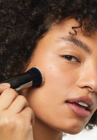 Make-upkwast met zachte haren, gehouden nabij de wang om product aan te brengen; model heeft krullend haar, een egale huid en een natuurlijke lipkleur.