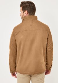 Braune Cordjacke mit hochgeschlossenem Kragen, gesteppten Schulterakzenten und geknöpften Manschetten. Schlichtes, strukturiertes Design ohne zusätzliche Verzierungen.