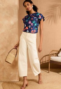 Ziedu raksta blūze tumši zila, ar viļņotu piedurknēm, sajaukta ar platām krēmkrāsas biksēm. Aksesuāri ietver austu somu un papēžu sandales.