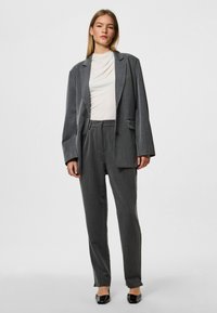 Grau maßgeschneiderter Anzug mit einem strukturierten Blazer und gefalteten Hosen; getragen über einem hellen Oberteil. Texturiertes Gewebe mit glatter Oberfläche.