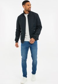 Svart bombarjacka med hög krage, två fickor fram, bärs över en grå skjorta och blå jeans, i kombination med vita sneakers.