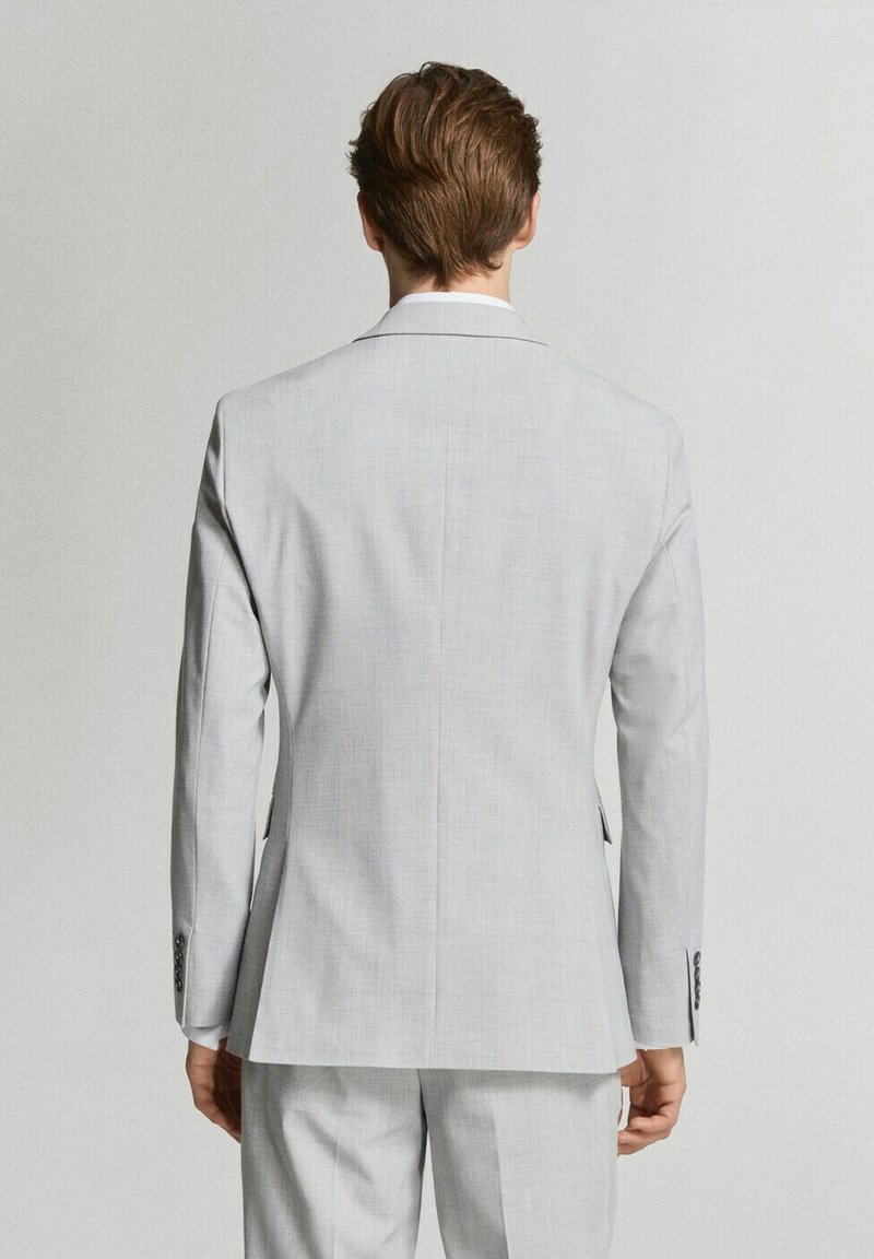 Man met kort bruin haar, gekleed in een lichtgrijs pak, gezien van achteren tegen een effen lichte achtergrond.