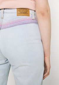 Vaaleansiniset farkut, joissa on vaaleanpunaisia ja violetteja raidallisia brodeerauksia takana, ja oikean taskun ylle on ommeltu vaalea nahkainen merkkilappu.