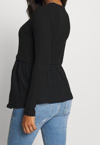Frau in einem schwarzen Langarmoberteil mit einem strukturierten schwarzen Peplum-Saum und hellblauen, ausgewaschenen Jeans, die vor einem schlichten, hellen Hintergrund steht.