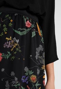 Tissu noir avec broderie florale colorée, présentant des motifs verts, jaunes, rouges et violets. Taille élastique et manches amples.