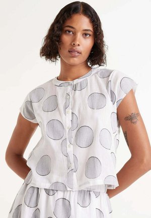Camisa blanca de manga corta con cuello redondo, con grandes círculos grises y cierre de botones en la parte delantera; tejido ligero y texturizado.