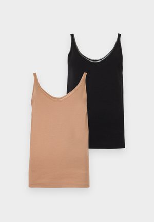 Kvinde står i neutral positur iført beige ærmeløs top, lang hvid nederdel og brune sandaler mod en ensfarvet hvid baggrund.