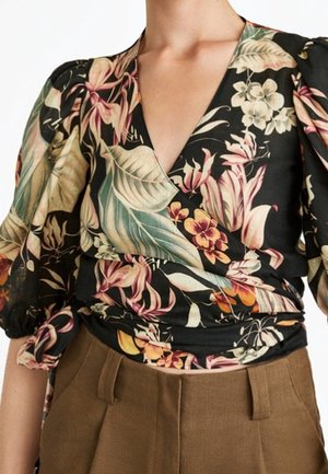 Kvinde iført en sort, blomstrede slå-om bluse med puffede ærmer, kombineret med højttaljede brune bukser.