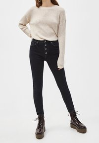 Crème ribbeltrui met brede mouwen, hoogewaisted zwarte skinny jeans met knoopsluiting, en glanzende bruine veterschoenen met plateauzolen.