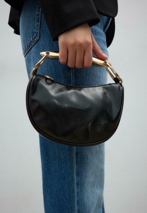 Roka drži majhno črno usnjeno torbico z ukrivljenim zlatim ročajem, oblečene v modre jeans hlače in vidno črno jakno.