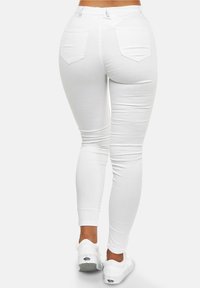 Vista trasera de una persona que lleva jeans blancos ajustados con bolsillos traseros y zapatillas blancas, de pie contra un fondo claro liso.