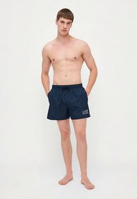 Νεαρός άνδρας που στέκεται ξυπόλητος, φορώντας σκούρα μπλε σορτς EA7 με τα χέρια στις τσέπες, μπροστά σε απλή φόντο.