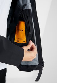 Main tenant une veste noire ouverte révélant une étiquette intérieure orange avec du texte et une petite poche zippée contre une doublure grise.