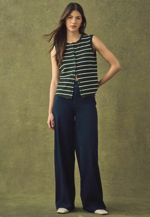 Zöld-fehér csíkos ujjatlan felső gombokkal, széles szárú sötétkék nadrággal és krémszínű cipővel, semleges háttéren.