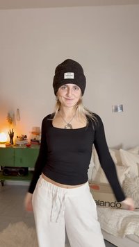 Μαύρο εφαρμοστό μακρυμάνικο τοπ, σε συνδυασμό με φαρδιά λευκά παντελόνια. Ένα σκούρο πλεκτό σκουφάκι έχει μια ετικέτα. Το φόντο περιλαμβάνει ένα πράσινο ντουλάπι και μαλακά έπιπλα.