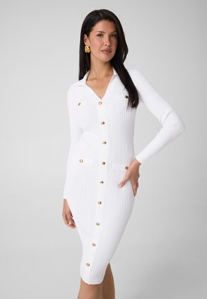 Femme aux longs cheveux foncés portant une robe blanche côtelée à boutons dorés et de grandes boucles d'oreilles dorées sur un fond clair uni.